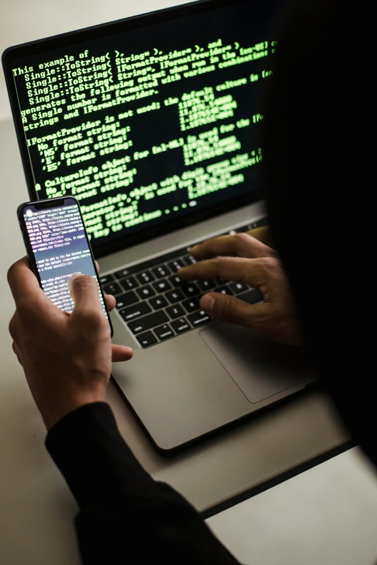 A hooded figure engaged in hacking using a laptop and smartphone in low light.